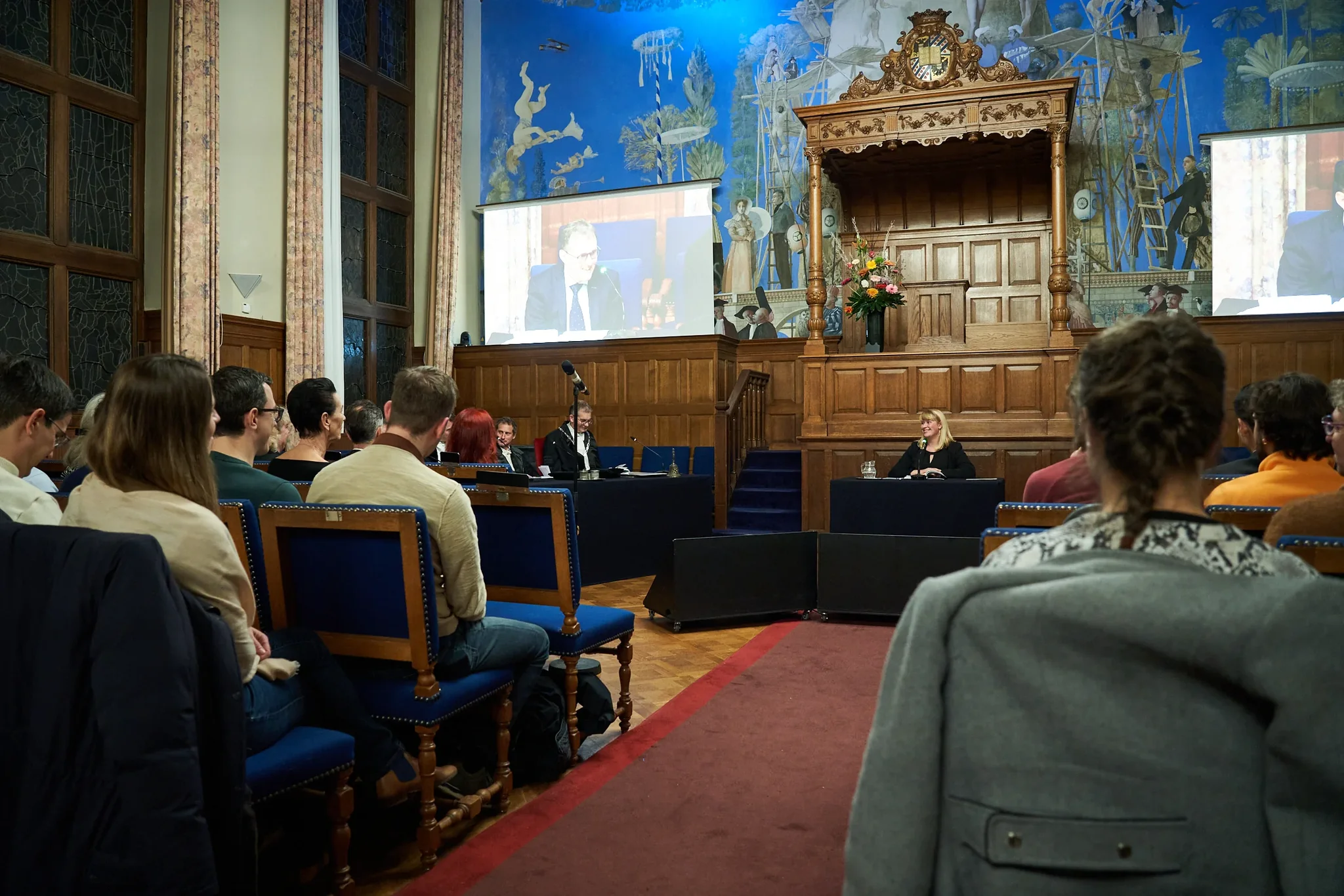 Promotieceremonie in een historische zaal met publiek en projectiescherm