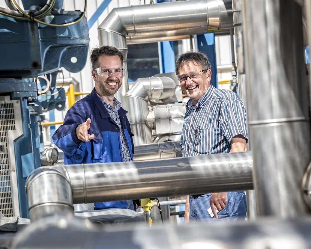Twee technici lachend naast industriële leidingen en machines