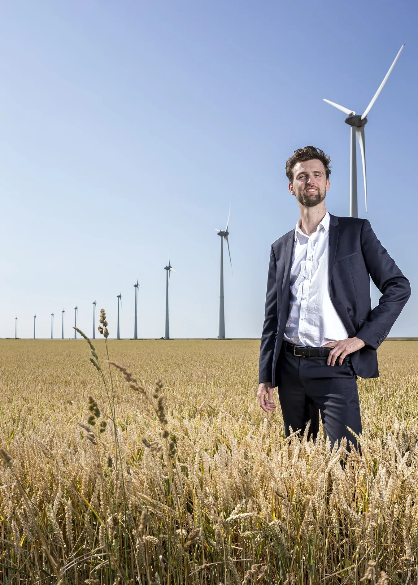 Man in blauw pak staat in een korenveld met windmolens op de achtergrond